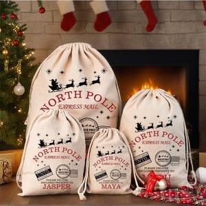 May include: A set of four beige canvas North Pole Express Mail drawstring gift bags. Each bag features a black graphic of Santa in his sleigh with reindeer, the words "North Pole Express Mail", and a name. The bags are in front of a fireplace.