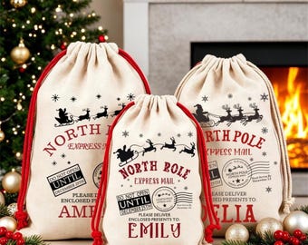May include: Three beige canvas drawstring gift bags with red cords. Each bag features black and red text reading North Pole Express Mail, a graphic of Santa in a sleigh, and a stamp stating Do Not Open Until December 25th. Names are printed at the bottom.