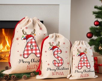 May include: Three beige drawstring gift bags made of fabric, each featuring a red and white buffalo plaid letter with reindeer antlers and a Santa hat. The bags are personalized with the names Allen, Sophia, and Allen, and the text Merry Christmas.