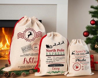 May include: Three beige drawstring gift bags with red and black text and graphics. The bags feature North Pole themed designs, including reindeer, sleighs, and personalized names Ashton, Carey, and Phoebe, with various postal-style stamps.