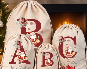 May include: Four beige drawstring gift sacks made of a linen-like fabric, each featuring a large red letter, a floral arrangement, and a cartoon Santa Claus. The sacks are personalized with names including Allen and Bella, and the text Merry Christmas.