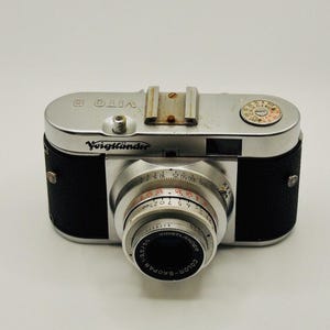 Pode incluir: Uma câmera Voigtländer Vito B prateada com uma cobertura de couro preto. A câmera tem uma lente prateada com um anel preto e um dial prateado com marcas vermelhas.