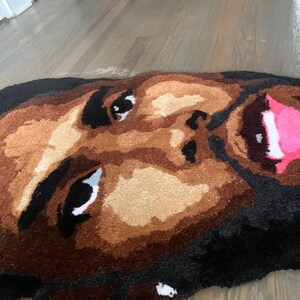 May include: A handmade rug featuring a detailed face design. The rug showcases a person's face with brown skin tones, black hair, and a pink mouth. The eyes are black and white. The rug is made of soft, textured material.