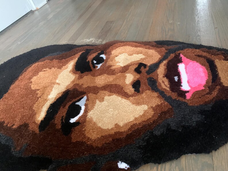 May include: A handmade rug featuring a detailed face design. The rug showcases a person's face with brown skin tones, black hair, and a pink mouth. The eyes are black and white. The rug is made of soft, textured material.