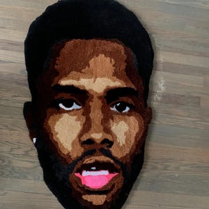 May include: A rug in the shape of a face, featuring a man with dark hair and a beard. The rug uses shades of brown, black, and pink to depict the facial features. The rug is placed on a wooden floor.