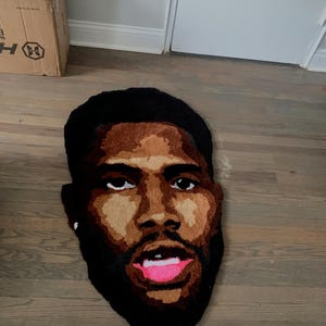 May include: A handmade rug in the shape of a face, featuring a dark-skinned person with a beard and open mouth. The rug has a black outline, brown skin tones, and pink lips. The rug is placed on a wooden floor.