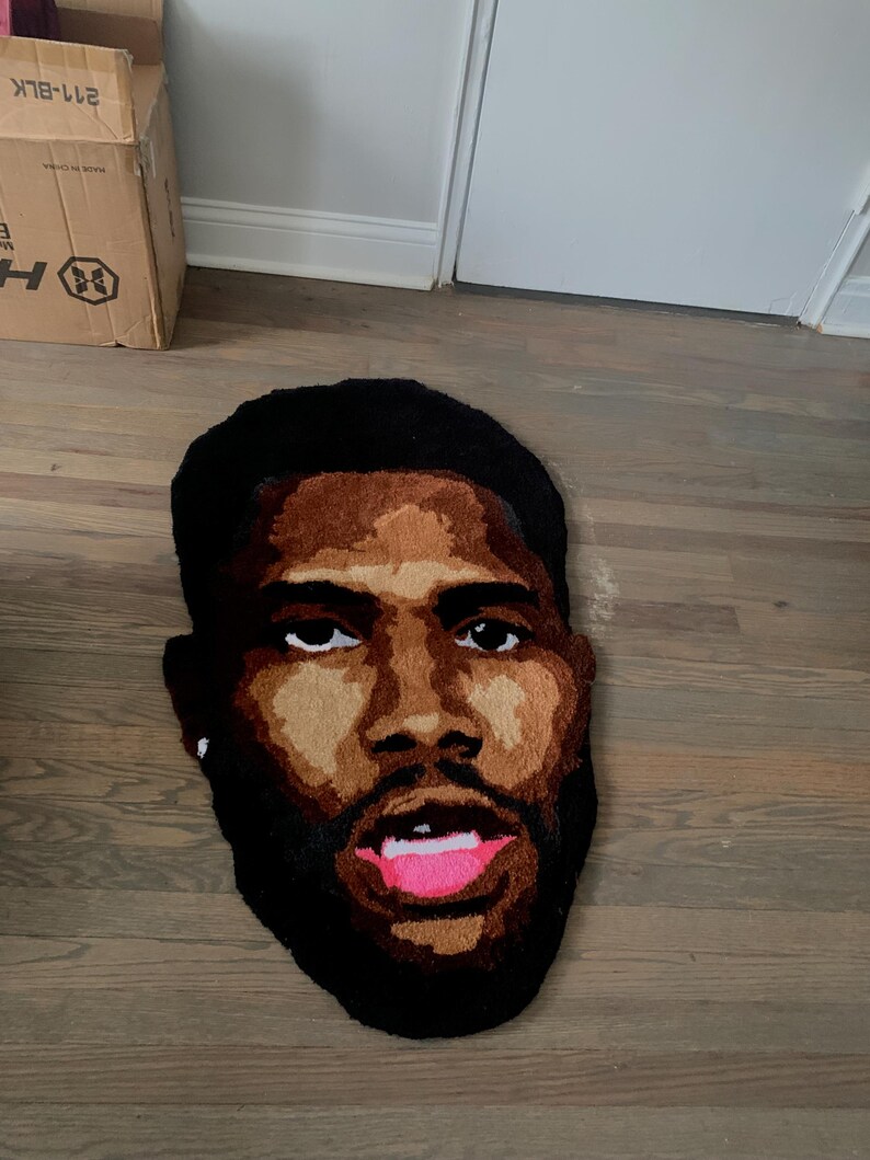 May include: A handmade rug in the shape of a face, featuring a dark-skinned person with a beard and open mouth. The rug has a black outline, brown skin tones, and pink lips. The rug is placed on a wooden floor.