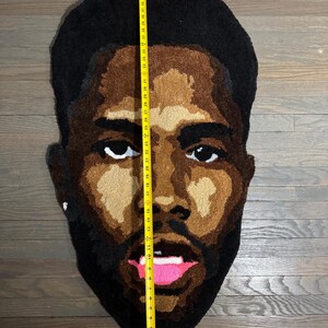 May include: A rug in the shape of a face, featuring a dark-skinned person with black hair and a beard. The rug is approximately 48 inches tall. The face has brown skin tones, white eyes, and a pink mouth.