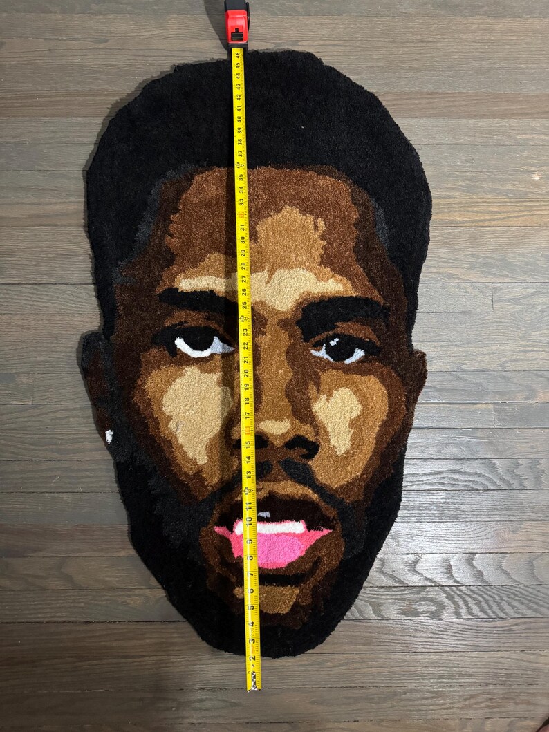 May include: A rug in the shape of a face, featuring a dark-skinned person with black hair and a beard. The rug is approximately 48 inches tall. The face has brown skin tones, white eyes, and a pink mouth.