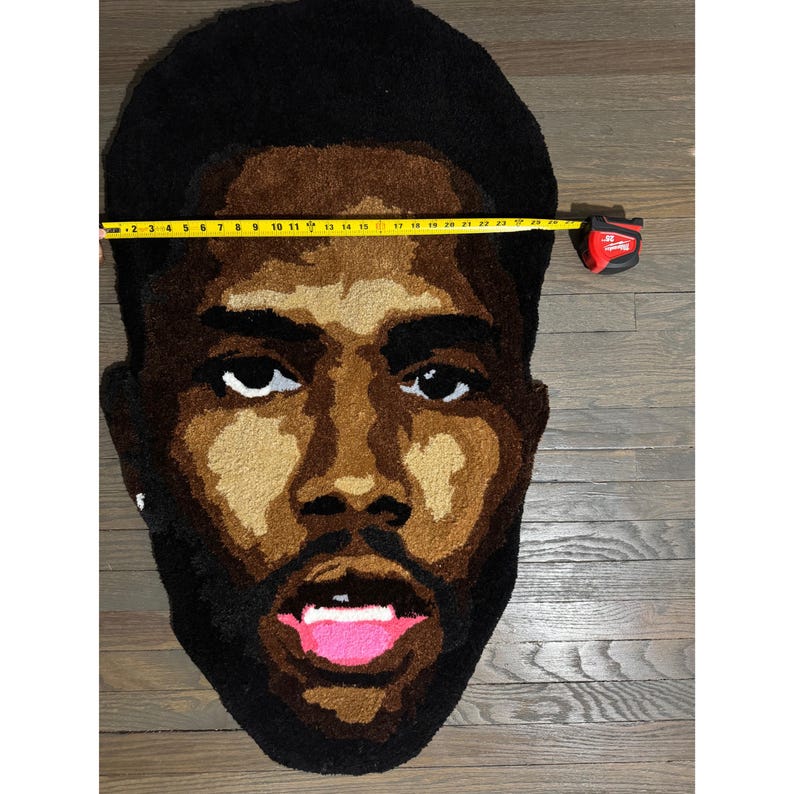 May include: A tufted rug in the shape of a man's face, featuring a dark brown afro, a light brown face, and a dark brown beard. The rug is approximately 23 inches wide. The mouth is open, revealing a pink tongue.