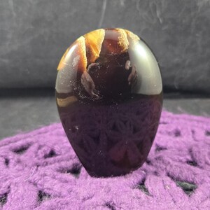 May include: A smooth, polished, dark brown and gold agate stone with a unique pattern. The stone is shaped like an egg and is sitting on a purple crocheted surface.