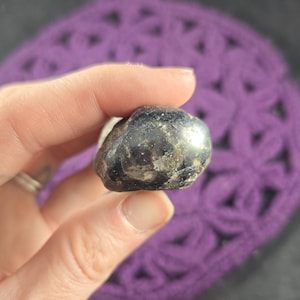 May include: A dark, polished stone with a reflective surface is held in a hand. The stone has a mottled appearance with areas of lighter color. The background is a purple crocheted item.