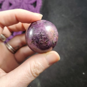May include: A polished, spherical gemstone in shades of deep purple, with areas of lighter pink and black inclusions. The crystal ball is held in a hand, with a blurred purple object in the background. The gemstone is approximately 2 inches in diameter.