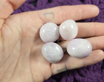 May include: Four smooth, white tumbled stones with a hint of lavender. The stones are in a hand, and the background is a purple knitted fabric.