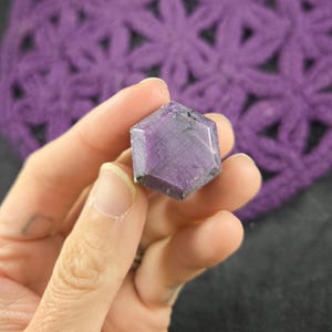 May include: A hand holds a faceted, hexagonal purple gemstone. The stone has a polished surface and a deep, rich colour. The background features a crocheted purple fabric with a floral pattern.