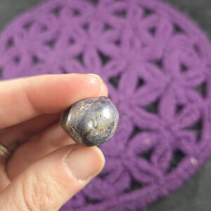 May include: A small, rounded, polished gemstone with a mix of gray, blue, and tan hues. The stone is held against a backdrop of a purple, open-weave fabric.