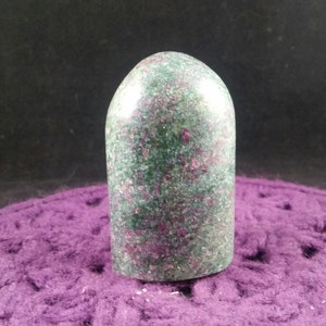 May include: A green and pink polished gemstone with a rounded top and a flat base. The gemstone is a natural crystal and has a unique, speckled appearance.
