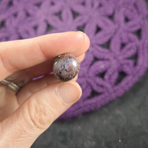 May include: A close-up of a round, iridescent purple and silver gemstone. The gemstone has a smooth, polished surface and is held in a hand.