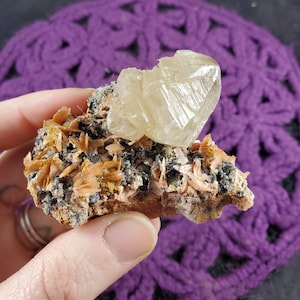 May include: A cluster of light brown and orange crystals on a dark gray rock with a large, clear, white crystal on top.