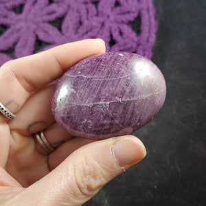 May include: A smooth, polished purple and white gemstone with a rounded shape. The stone has a striated pattern and a glossy finish.