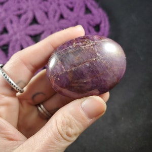 May include: A polished purple and red gemstone with a smooth, oval shape. The stone has a striated pattern and a glossy finish.