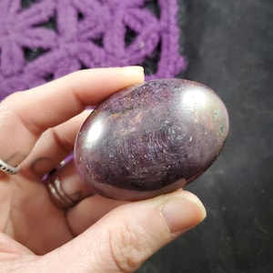 May include: A smooth, polished purple and grey gemstone with a rounded, oval shape. The stone is held in a hand.