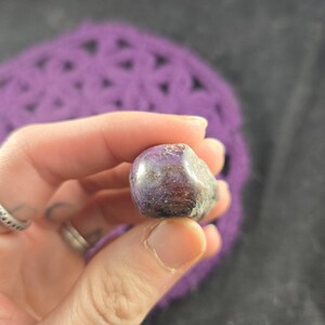 K&ouml;nnte beinhalten: Ein polierter, abgerundeter Stein mit einer Mischung aus lila, braunen und grauen Farbt&ouml;nen. Der Stein wird in einer Hand gehalten, mit einem verschwommenen lila Hintergrund. Die Oberfl&auml;che des Steins erscheint glatt und reflektierend, mit Farbvariationen.