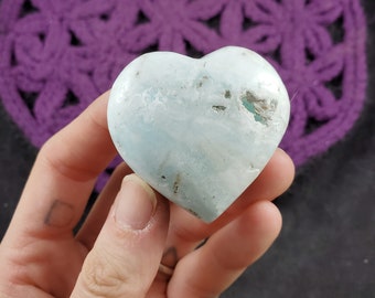 May include: A light blue heart-shaped crystal with white and grey inclusions. The crystal is smooth and polished.