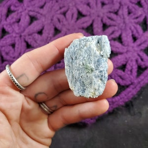 Peut inclure: Un cristal de kyanite bleu avec une surface rugueuse et texturée. Le cristal est de couleur bleu clair avec quelques inclusions bleu foncé et vertes.