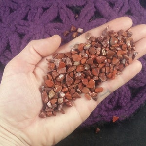 May include: A close-up of a hand holding a pile of small, red, brown, and white tumbled stones. The stones are irregularly shaped and have a rough texture.