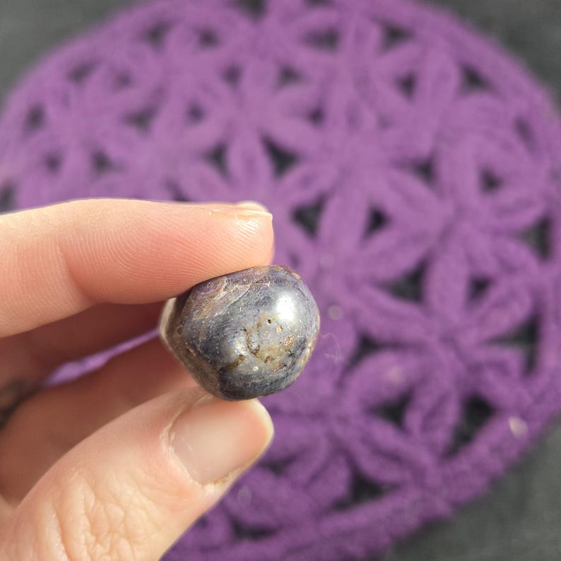 May include: A small, polished, dark blue gemstone with brown and tan inclusions. The stone is held between fingers against a blurred background of a purple, open-weave fabric.