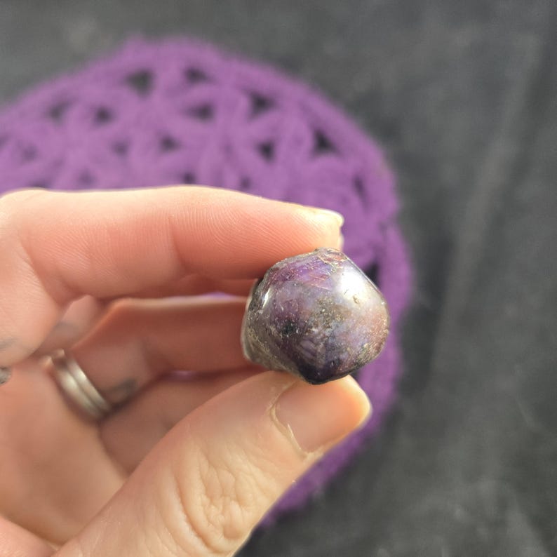 K&ouml;nnte beinhalten: Ein polierter, abgerundeter Edelstein mit tiefvioletter Farbe, gesprenkelt mit helleren Farbt&ouml;nen und Goldt&ouml;nen. Der Stein wird in einer Hand gehalten, mit einem verschwommenen lila Hintergrund.