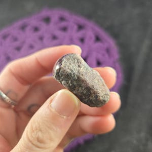 K&ouml;nnte beinhalten: Ein kleiner, rau strukturierter, grau-brauner Stein mit polierter, rot-violetter Oberseite. Der Stein wird zwischen den Fingern vor einem verschwommenen, violetten Hintergrund gehalten. Der Stein scheint ein nat&uuml;rliches Mineral oder Edelstein zu sein.