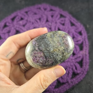 May include: A polished, oval-shaped stone with a mix of colours including purple, grey, and green. The stone is held in a hand, showcasing its smooth texture and natural patterns. The background features a purple crocheted item.