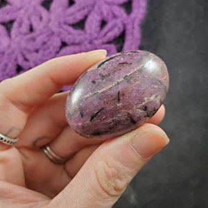 May include: A smooth, polished purple and black gemstone with a swirling pattern. The gemstone is shaped like an oval and is held in a hand.