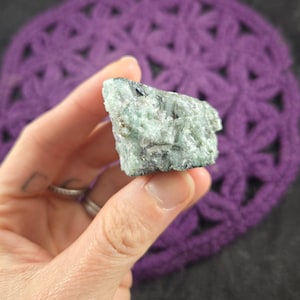 May include: A rough, green emerald crystal with black inclusions. The crystal is about the size of a small egg.