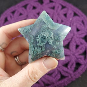 May include: A green and gray moss agate star-shaped crystal. The crystal has a natural, rough texture and is translucent.