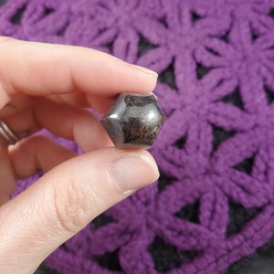 May include: A dark brown and black gemstone with a faceted, geometric shape. The gemstone is held in a hand.