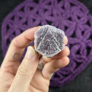 May include: A hexagonal raw crystal specimen with a deep purple and magenta hue. The stone features a textured, crystalline surface with visible natural fractures and a geometric, six-sided shape that tapers slightly toward the edges.