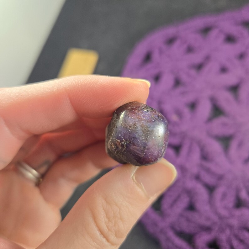K&ouml;nnte beinhalten: Ein polierter, abgerundeter Edelstein mit tiefvioletter Farbe, gesprenkelt mit helleren Farbt&ouml;nen und braunen Akzenten. Der Stein wird zwischen Fingern gehalten, wodurch seine glatte, reflektierende Oberfl&auml;che zur Geltung kommt. Im Hintergrund befindet sich ein lila geh&auml;keltes Objekt.