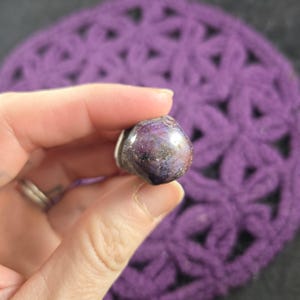 K&ouml;nnte beinhalten: Ein polierter, violetter Edelstein mit silbernem Sockel wird in einer Hand gehalten. Der Stein hat eine abgerundete Form und eine Mischung aus dunklen und hellen Lilat&ouml;nen. Der Hintergrund ist ein geh&auml;keltes Objekt in Lila.