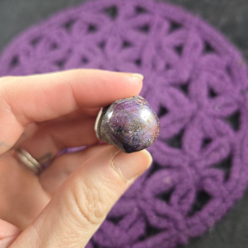 K&ouml;nnte beinhalten: Ein polierter, violetter Edelstein mit silbernem Sockel wird in einer Hand gehalten. Der Stein hat eine abgerundete Form und eine Mischung aus dunklen und hellen Lilat&ouml;nen. Der Hintergrund ist ein geh&auml;keltes Objekt in Lila.