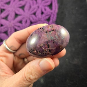 May include: A polished purple and black gemstone with a smooth, oval shape. The stone has a high gloss finish and is held in a hand.