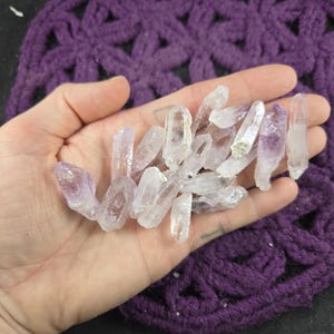 May include: A handful of small, clear amethyst crystal points with purple tips. The crystals are held in a hand, with a purple crocheted background.