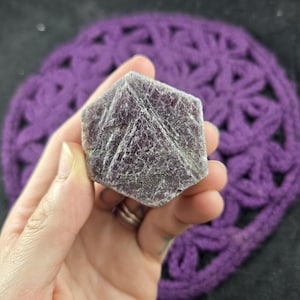 May include: A hand holds a dark purple, cube-shaped crystal with a textured surface and white striations. The crystal is set against a blurred background of a purple, crocheted item.
