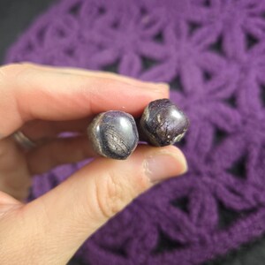 May include: Two small, tumbled, dark purple and grey stones with a smooth, polished surface and visible natural inclusions. The stones have an irregular, rounded shape and exhibit a subtle sheen across their textured, multi-toned surfaces.