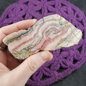 May include: A pink and grey banded, polished, natural stone with a rough, irregular edge. The stone has a swirling pattern of pink, grey, and white.
