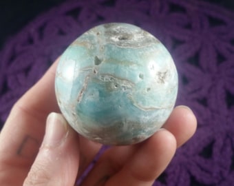 May include: A polished sphere of blue-green aragonite, featuring swirling patterns of light brown and gray. The gemstone is held in a hand, showcasing its smooth, rounded form. The sphere's surface has natural variations in color and texture.