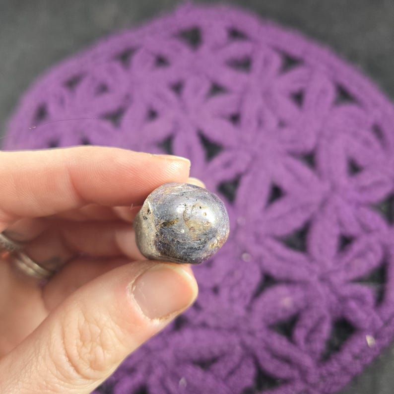 May include: A small, rounded, polished stone with a dark, metallic gray color and subtle variations. The stone is held in a hand, with a purple crocheted background. The stone appears to be a gemstone or decorative item.