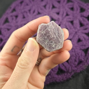 May include: A rough, pink-red ruby crystal with a hexagonal shape. The crystal has a natural, unpolished finish and is held in a hand.